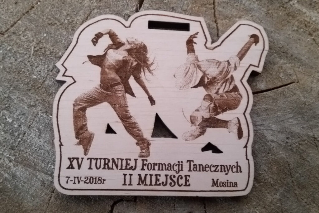 Nieregularnych kształtów drewniany medal z XV Turnieju Formacji Tanecznych w Mosinie, ozdobiony grawerem przedstawiającym sylwetkę tancerek w dynamicznej pozie.