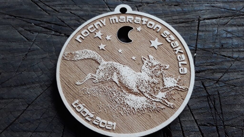 Okrągły drewniany medal na Nocny Maraton Szakala, ozdobiony precyzyjnym grawerem wyjącego szakala na tle lasu i nocnego nieba z księżycem.