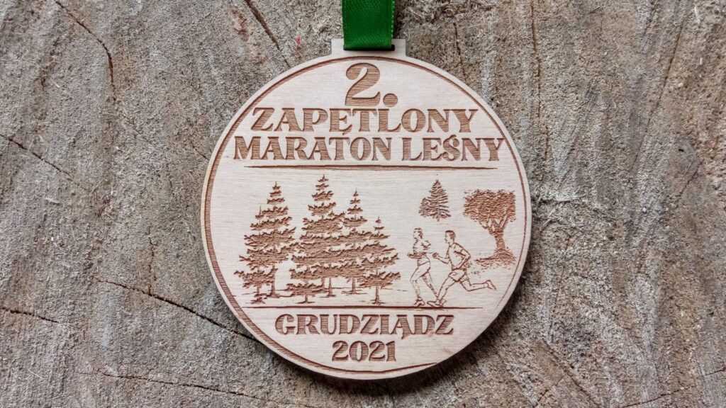 Okrągły drewniany medal na Zapętlony Maraton Leśny, przedstawiający grawerowaną ścieżkę biegnącą przez las iglasty z centralnie umieszczoną nazwą biegu i biegaczami.