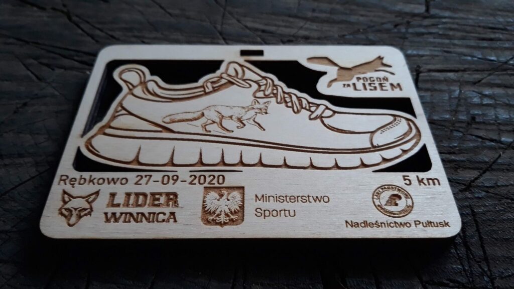 Prostokątny drewniany medal na bieg leśny z ażurowym wycięciem w kształcie buta biegowego, na dole logotypy.