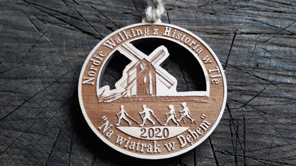 Tekst alternatywny (Alt):
Ażurowy drewniany medal na rajd Nordic Walking „Z Historią w Tle” z precyzyjnym grawerem zabytkowego wiatraka i sylwetkami maszerujących osób.