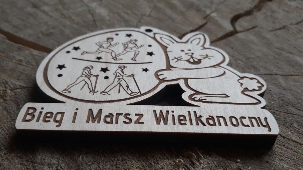 Oryginalny drewniany medal na Bieg i Marsz Wielkanocny z grawerem zająca trzymającego pisankę, na pisance wygrawerowani biegacze i chodziarze, oraz wycięte ażurowe gwiazdki, na dole napis Bieg i Marsz Wielkanocny.