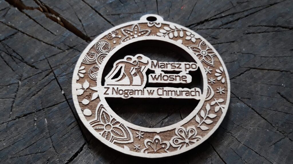 Wiosenna ramka medalu a w niej ażurowo wycięci chodziarze, czyli drewniany medal na „Marsz po Wiosnę”. Wiosenna ramka medalu a w niej ażurowo wycięci chodziarze, czyli drewniany medal na „Marsz po Wiosnę”.