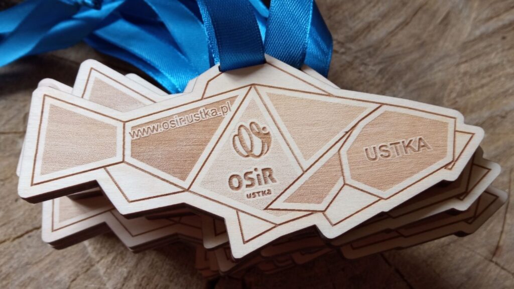 Drewniany medal wycięty w realistycznym kształcie modernistycznej ryby z napisami i logiem, zawieszony na niebieskiej wstążce.