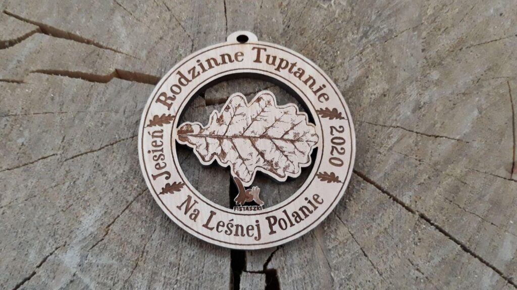 Okrągły drewniany medal z wydarzenia „Rodzinne Tuptanie”, posiadający ażurowe wycięcie w kształcie liścia dębu. Wokół napis: Rodzinne Tuptanie na Leśnej Polanie.