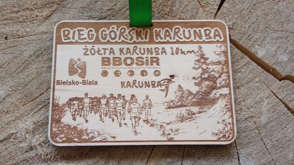 Prostokątny drewniany medal na „Bieg Górski Karunba” o solidnej grubości 6 mm, z precyzyjnym grawerem zawodników biegnących po nierównym, górzystym terenie w otoczeniu leśnej roślinności. Medal na żółtej zielonej, na medalu grawer 3 logotypów sponsorów.