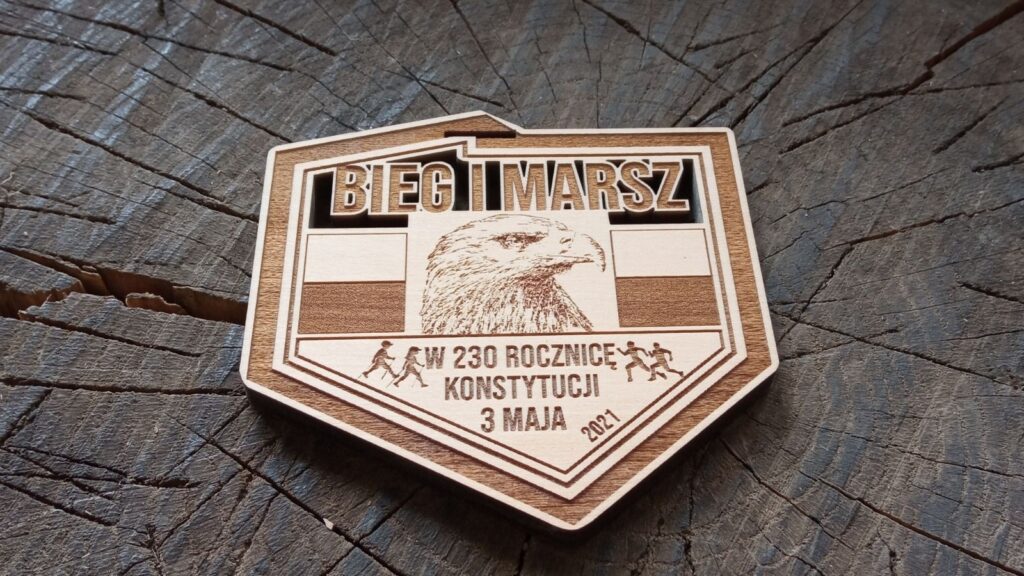 Drewniany medal o grubości 6 mm w kształcie konturu Polski na „Bieg i Marsz w 230. Rocznicę Konstytucji 3 Maja”, z grawerowanym orłem, biało-czerwoną flagą oraz okolicznościowymi napisami.