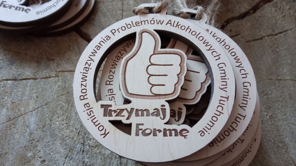 Okrągły drewniany medal ażurowy z wyciętym motywem „kciuka w górę” wewnątrz koła oraz grawerowanym hasłem „Trzymaj formę!” i nazwą Gminnej Komisji Rozwiązywania Problemów Alkoholowych.
