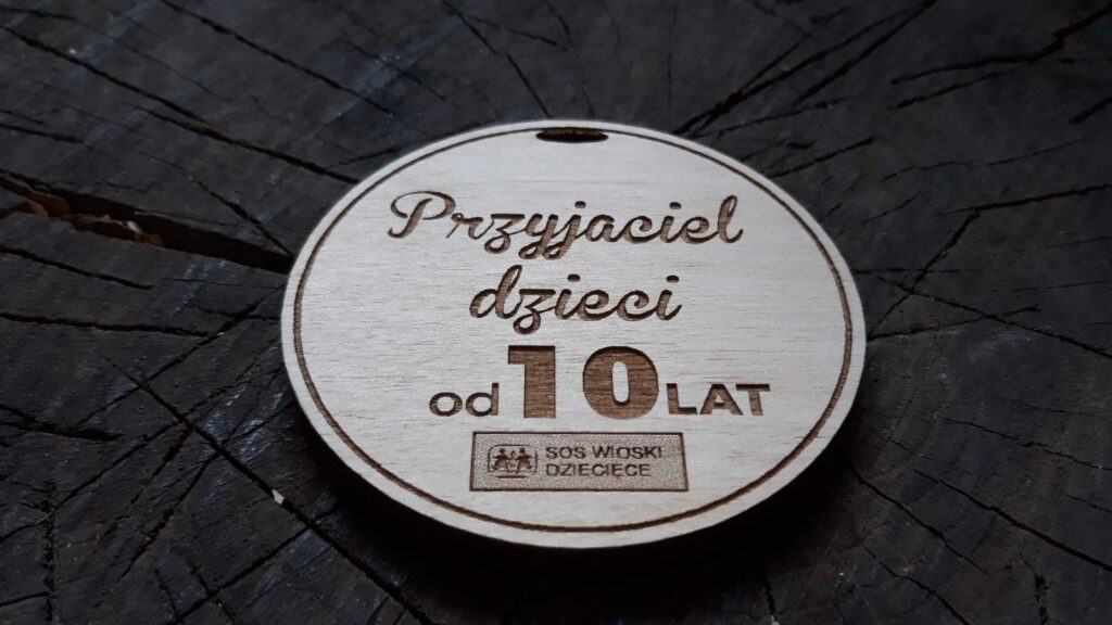 Drewniany medal pamiątkowy z napisem „Przyjaciel Dzieci”, ozdobiony grawerem o logotypem SOS Wioski Dziecięce.