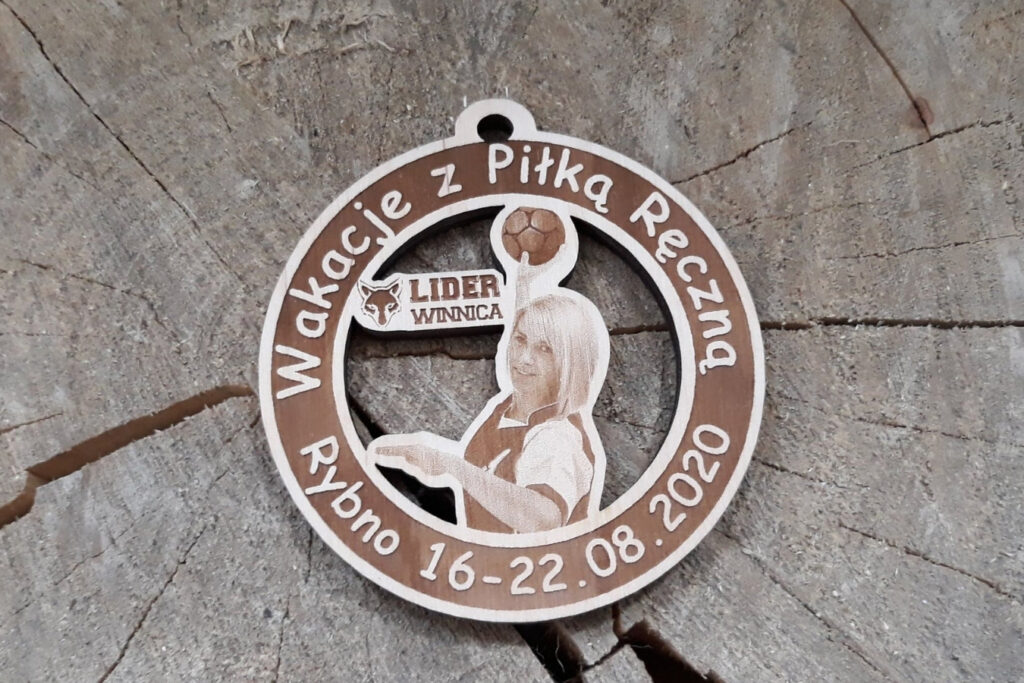 Okrągły drewniany medal na „Wakacje z Piłką Ręczną” z ażurowo wyciętą sylwetką szczypiornistki i logotypem.