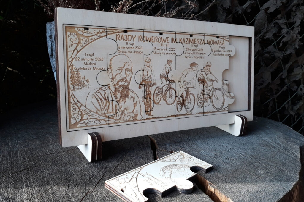 Zestaw sześciu drewnianych medali modułowych na rajd rowerowy, które po złożeniu tworzą prostokątną tabliczkę z grawerem kolarzy na trasie, logotypem wydarzenia i mapą, wykonany z jasnej brzozy bez śladów osmaleń. medale zamieszczone w drewnianej ramce