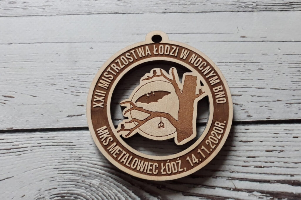 Ażurowy drewniany medal na Mistrzostwa w Nocnym Biegu Na Orientację z precyzyjnym wycięciem symbolĻw nocnych - drzewa, nietoperza