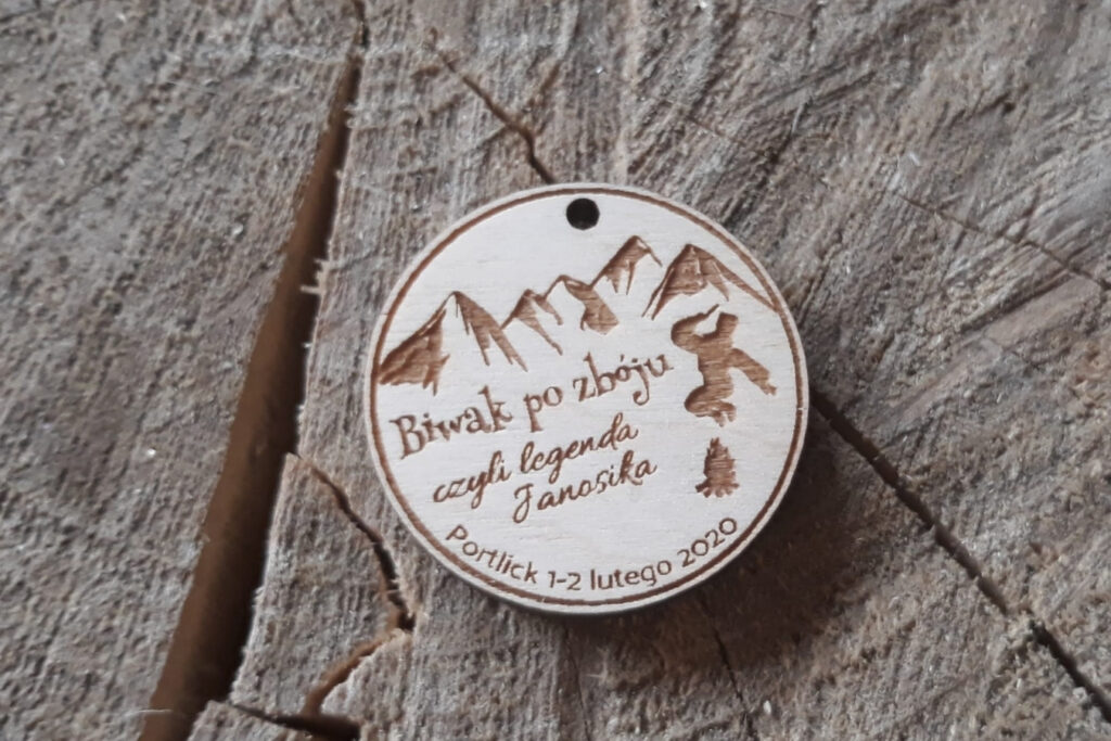 Tekst alternatywny (Alt):
Okrągły drewniany medal pamiątkowy z grawerem przedstawiającym biwak górski: namiot rozstawiony u podnóża strzelistych szczytów.