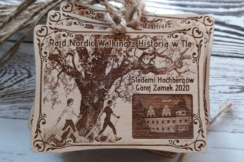 Prostokątny drewniany medal na rajd Nordic Walking z grawerem historycznego zamku w Goraju, otoczony motywami leśnymi i sylwetkami maszerujących osób.