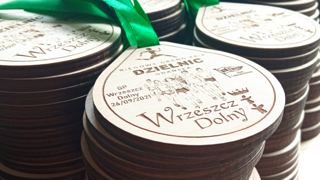Owalny drewniany medal Grand Prix Dzielnic Gdańska – etap Wrzeszcz Dolny, z grawerem biegaczy oraz oficjalnymi logotypami organizatorów.