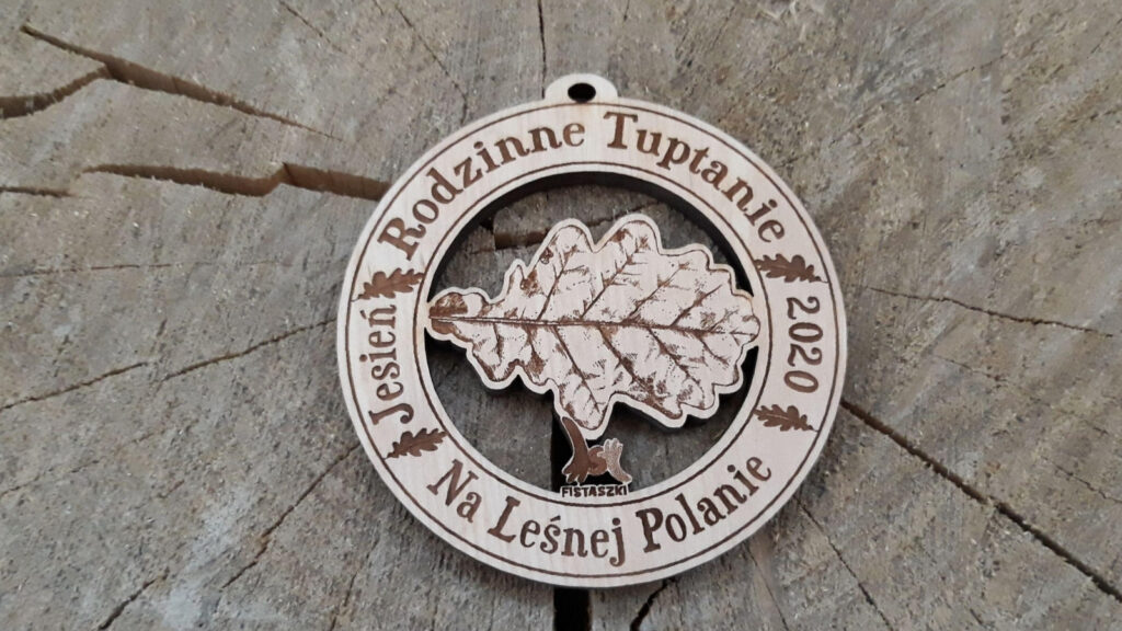 Okrągły drewniany medal z wydarzenia „Rodzinne Tuptanie”, posiadający ażurowe wycięcie w kształcie liścia dębu. Wokół napis: Rodzinne Tuptanie na Leśnej Polanie.