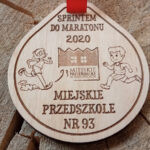 Drewniany medal o solidnej grubości 6 mm na zawody dla dzieci „Sprintem do maratonu 2020”, z precyzyjnym grawerem uśmiechniętych, biegnących postaci dzieci, gwiazdek oraz motywujących napisów, dedykowany najmłodszym sportowcom.
