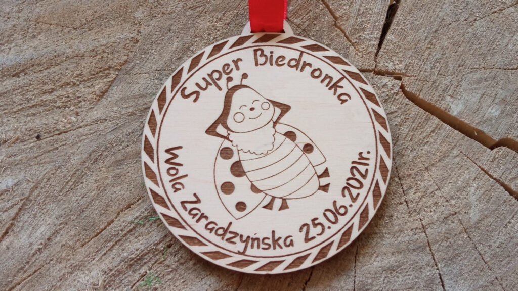 Okrągły drewniany medal dla dzieci na zakończenie przedszkola z grawerem uśmiechniętej biedronki w pelerynie superbohatera oraz napisem „Super Biedronka”.