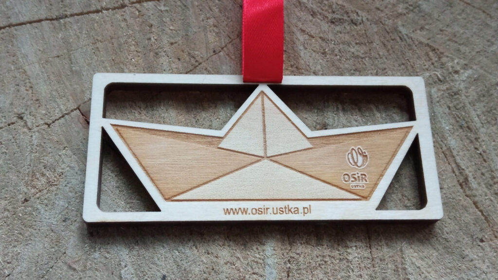 Ażurowy drewniany medal o solidnej grubości 6 mm, z misternie wyciętym na wylot motywem żaglówki (łódki)  oraz zawieszoną kontrastującą, elegancką czerwoną wstążką.