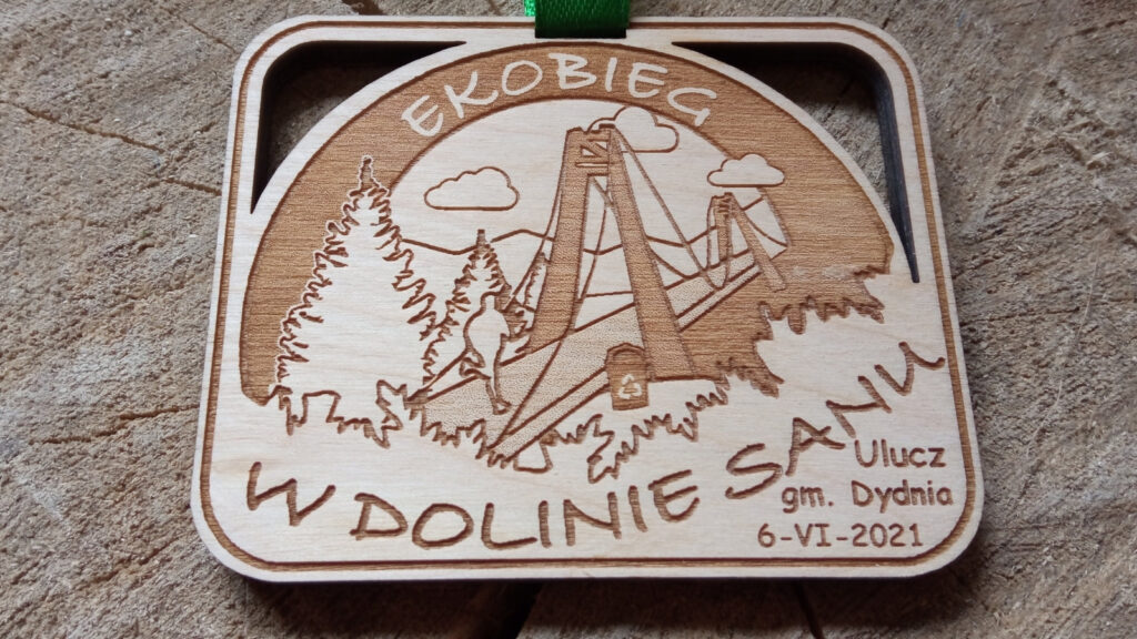 Prostokątny drewniany medal o grubości 6 mm na „Ekobieg w Dolinie Sanu” z wygrawerowanym motywem rzek i mostu, oraz pobliskich drzew. Na most wbega biegacz, góra mostu wycięta ażurowo, całosć na zielonej wstążce.