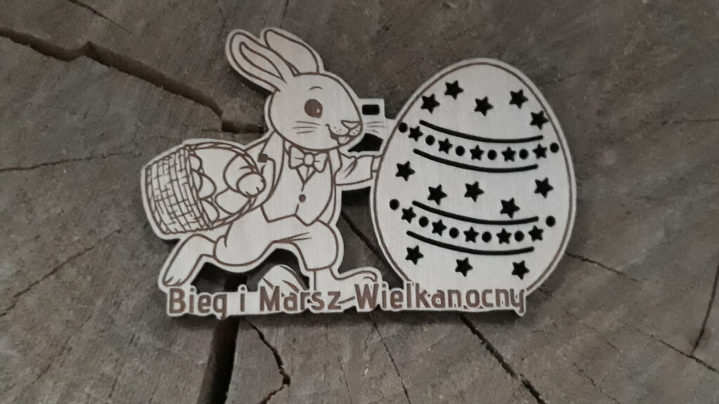 Oryginalny drewniany medal na Bieg i Marsz Wielkanocny z grawerem zająca trzymającego pisankę, na pisance wygrawerowani biegacze i chodziarze, oraz wycięte ażurowe gwiazdki, na dole napis Bieg i Marsz Wielkanocny.