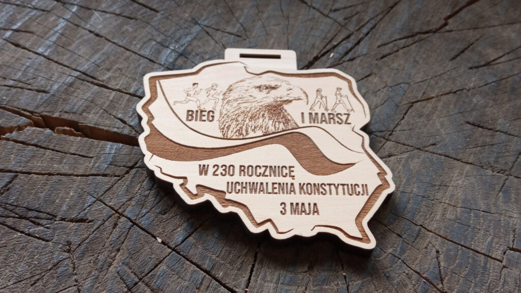 Patriotyczny drewniany medal o solidnej grubości 6 mm na „Bieg i Marsz w 230. Rocznicę Konstytucji 3 Maja”, z precyzyjnym grawerem Orła Białego w koronie, stylizowaną historyczną czcionką oraz motywem gałązek dębowych symbolizujących trwałość i pamięć.