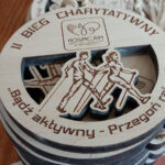 Okrągły ażurowy drewniany medal o solidnej grubości 6 mm na „II Bieg Charytatywny”, z misternie wyciętym na wylot symbolem splecionych kijków Nordic Walking, motywem wstążki onkologicznej oraz głębokim grawerem hasła „Bądź aktywny – przegoń raka!”.