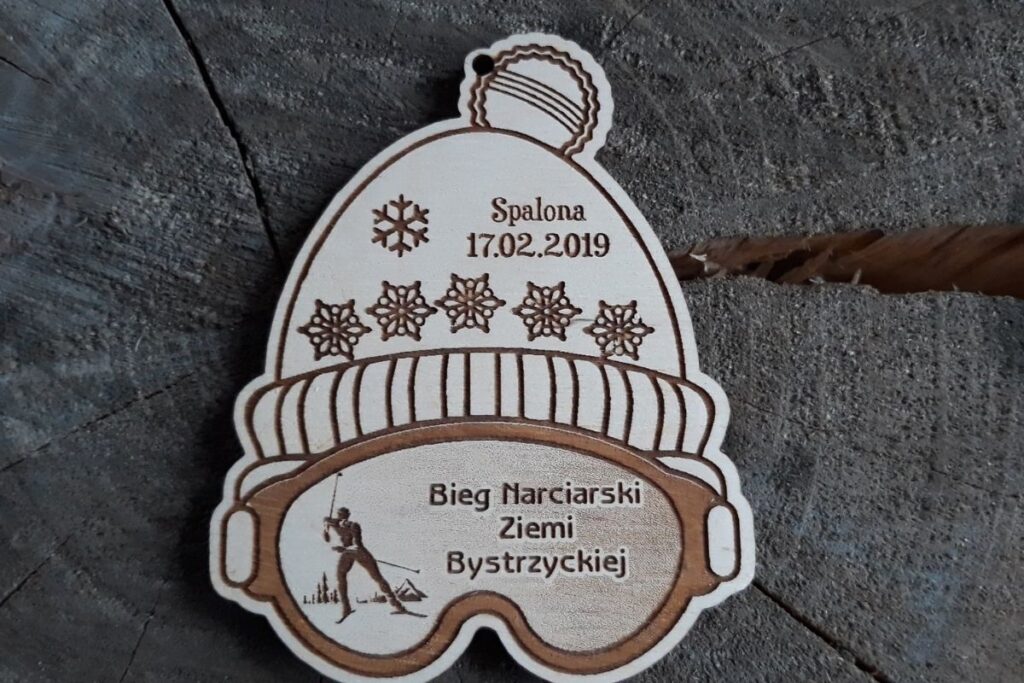 Drewniany medal pamiątkowy z Biegu Narciarskiego Ziemi Bystrzyckiej 2019 w Spalonej z grawerem narciarza. Medal w kształcie czapki z narciarskimi googlami.