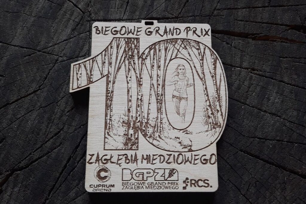Propozycja drewnianego medalu na 10. Biegowe Grand prix Zagłębia Miedziowego. Drewniany medal z cyfrą 10 napisami i logiem.