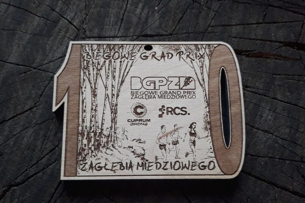 Propozycja drewnianego medalu na 10. Biegowe Grand prix Zagłębia Miedziowego. Drewniany medal z cyfrą 10 napisami i logiem.