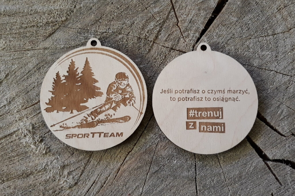 Okrągły drewniany medal z grawerem narciarza w dynamicznym zjeździe, z motywującym cytatem.