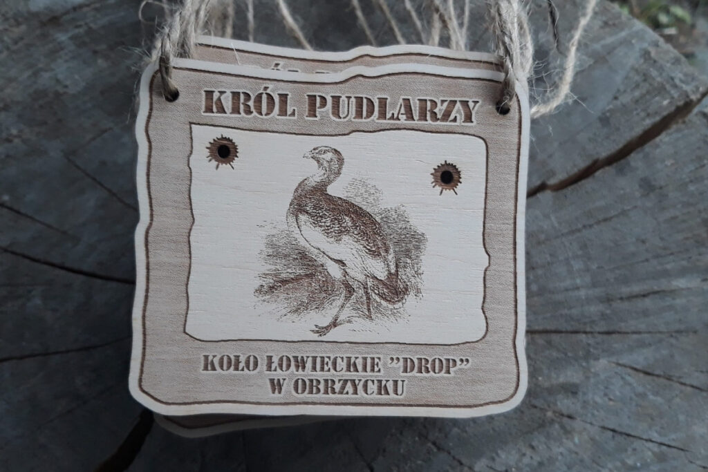 Prostokątny drewniany medal z grawerem dropia, napisem Król Pudlarzy oraz kulami - wycięciami na wylot.