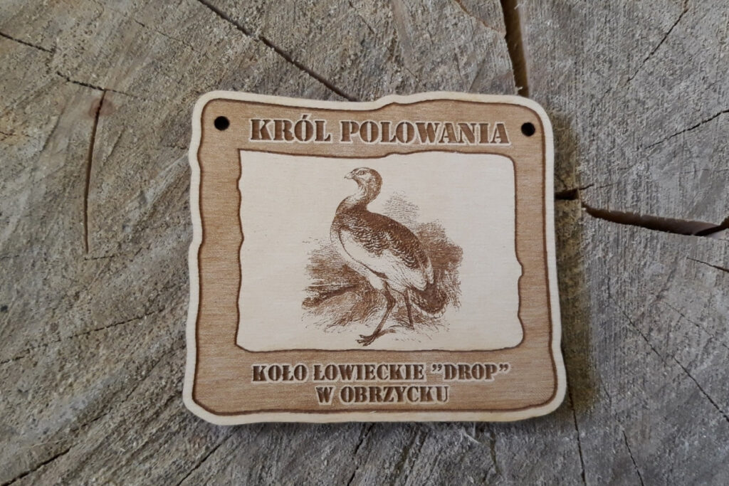 Prostokątny drewniany medal z grawerem dropia w dumnej postawie, napisem Król Polowania oraz datą wydarzenia.
