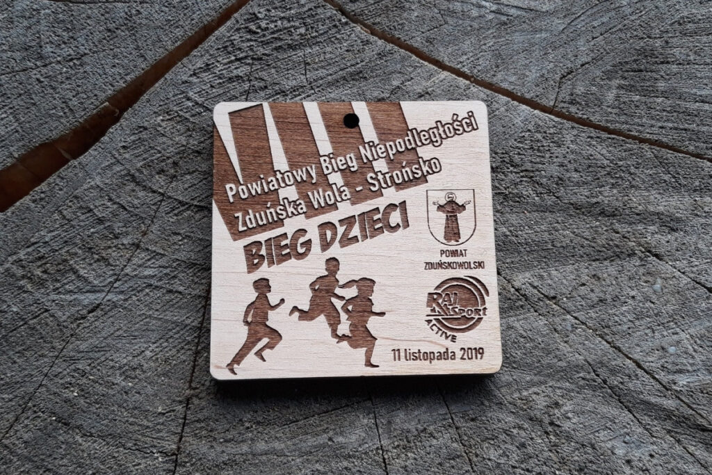 Drewniany, kwadratowy medal z napisami: Powiatowy Bieg Niepodległości - VII Bieg Dzieci, logotypami i biegnącymi dziećmi.