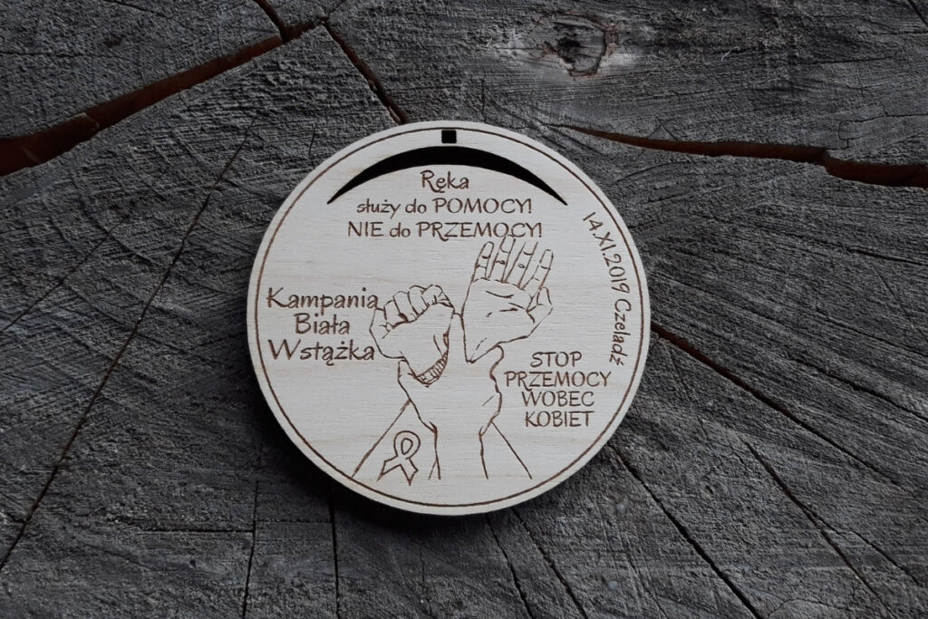 Okrągły drewniany medal z grawerem splecionych dłoni, napisem Stop Przemocy Wobec Kobiet oraz symbolem białej wstążki.