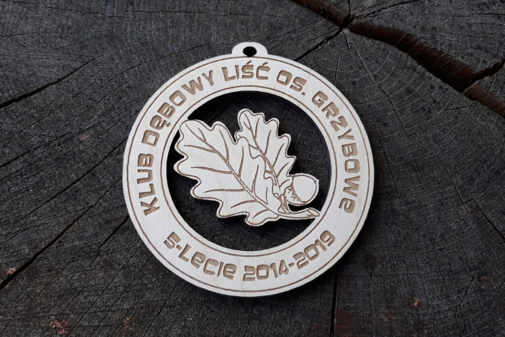 Okrągły drewniany medal z ażurowym wycięciem w kształcie liścia dębu, wykonany z jasnej brzozy, z grawerowanymi napisami po okręgu.