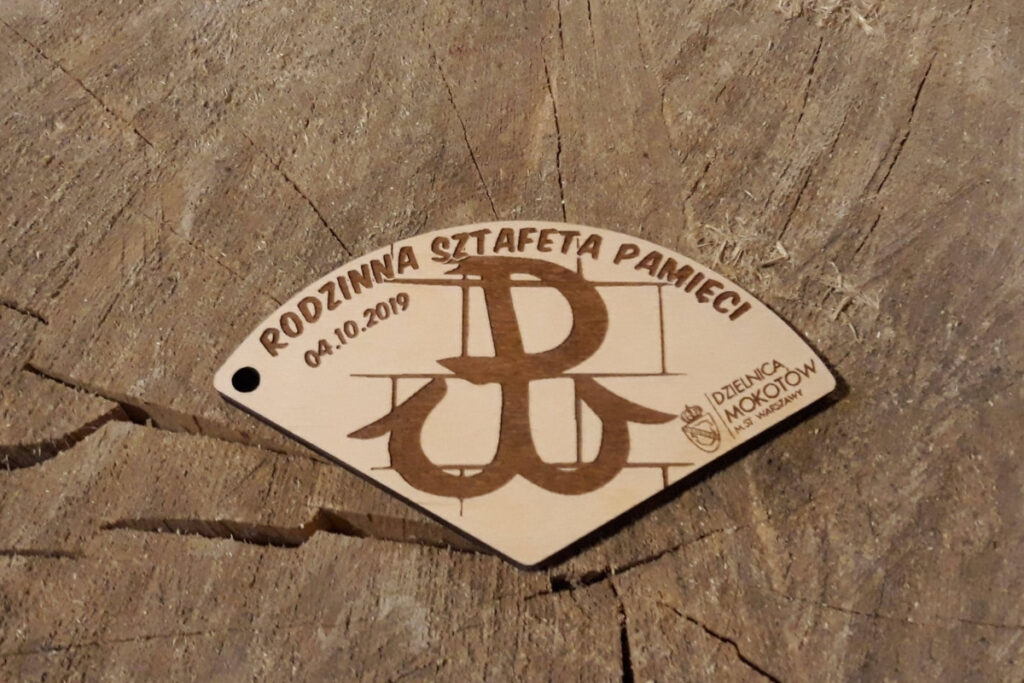 Trzy drewniane medale w kształcie wycinków koła, które po złączeniu tworzą kompletną okrągłą tabliczkę z grawerem Rodzinna Sztafeta Pamięci.