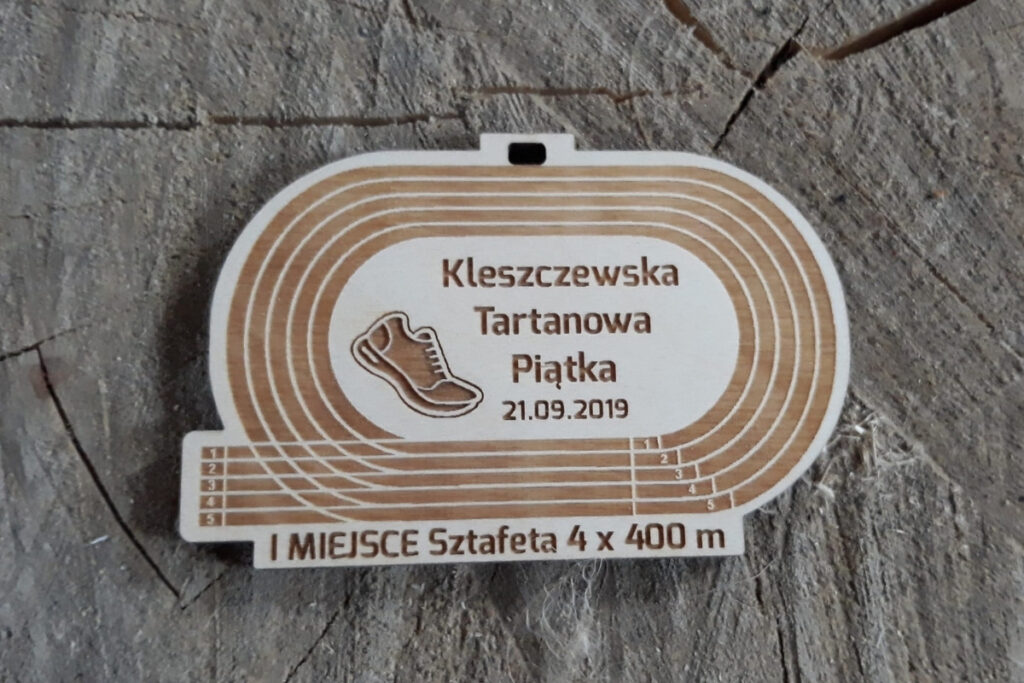 Drewniany medal w kształcie owalnej bieżni lekkoatletycznej z wygrawerowanymi torami, napisem oraz grafiką buta.