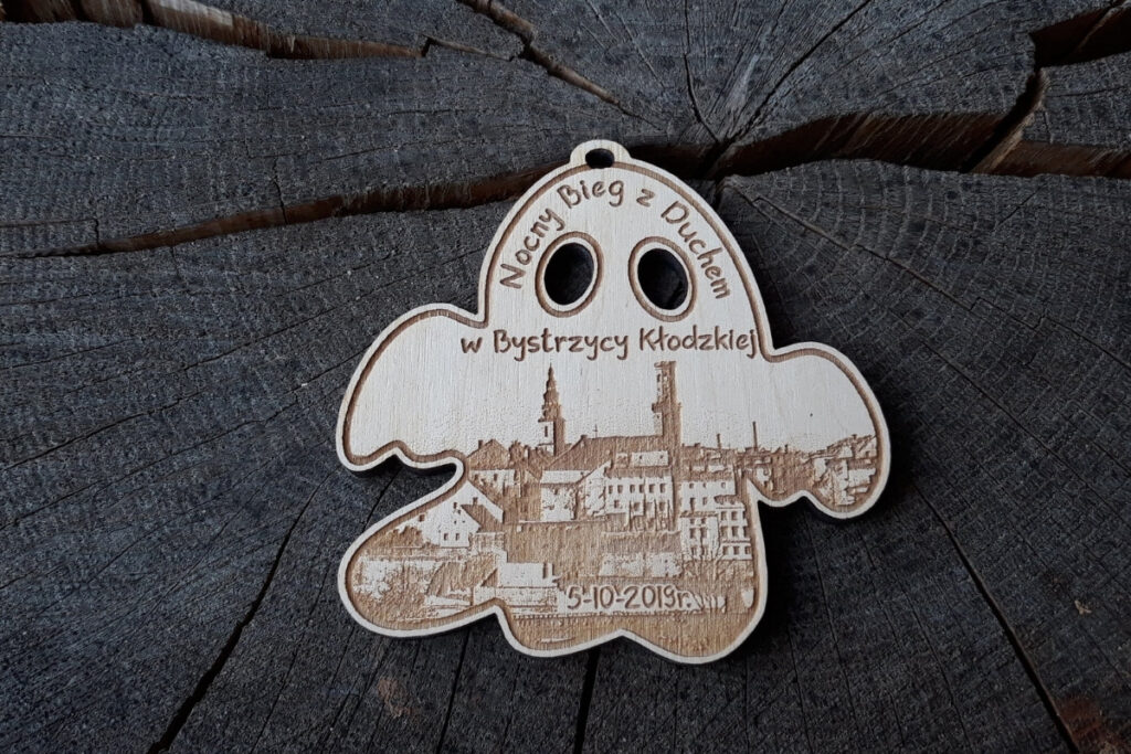 Drewniany medal wycięty w kształcie  ducha z grawerem Nocny Bieg z Duchem, rycin miasta i wyciętymi ażurowo na wylot oczami.