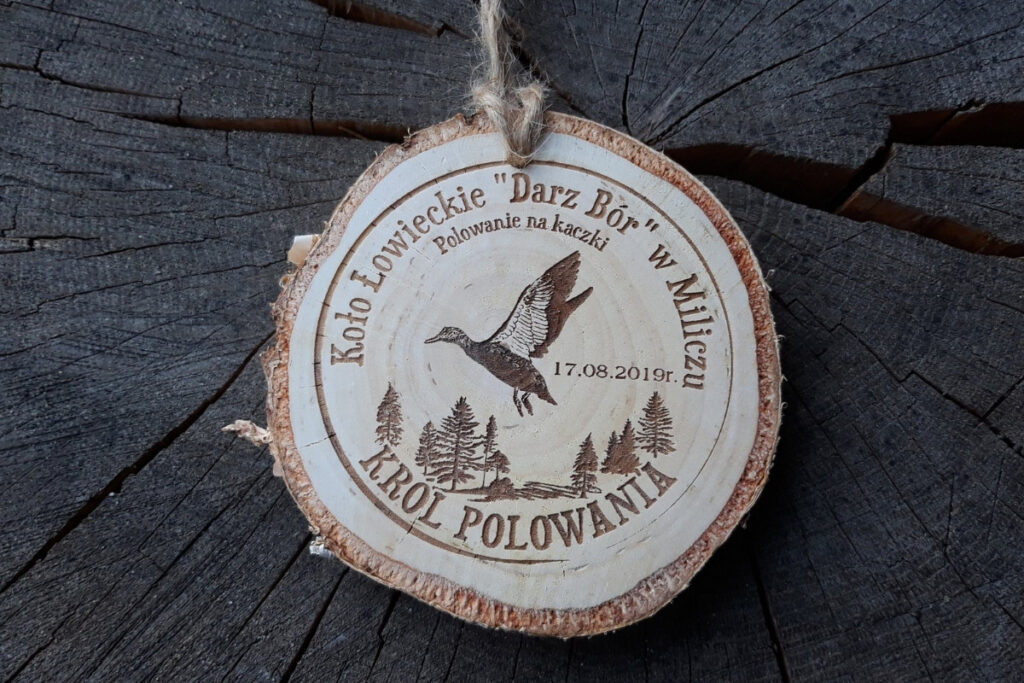 Medal z naturalnego plastra brzozy z korą, z grawerem kaczki krzyżówki zrywającej się do lotu, napisem Polowanie na Kaczki i ozdobnym motywem zalesienia.