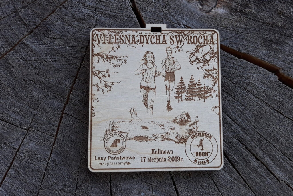 Kwadratowy medal z drewna brzozowego z grawerem gęstego lasu, sylwetką biegacza i napisem Leśna Dycha.
