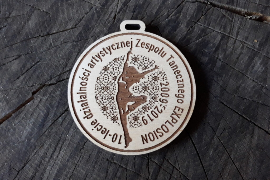Okrągły drewniany medal z grawerem logotypu zespołu artystycznego, napisem 10-lecie oraz tancerką.