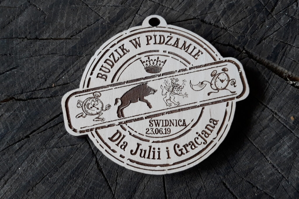 Okrągły drewniany medal z grawerem budzika, napisem Bieg Budzika dla Julii i Gracjana oraz datą wydarzenia.
