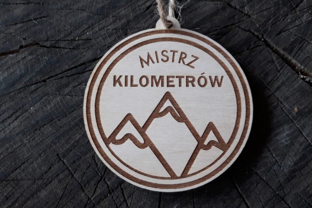 Okrągły drewniany medal z grawerem uskrzydlonego buta biegowego, wieńcem laurowym i napisem Mistrz Kilometrów.