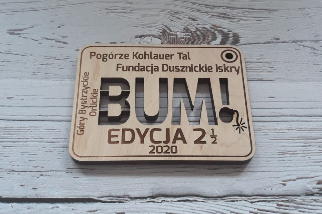 Prostokątny drewniany medal z grawerem stylizowanej, komiksowej bomby z zapalonym lontem i napisem wyciętym ażurowo „BUM”, wykonany z czystej brzozy bez śladów przypaleń i dymu.