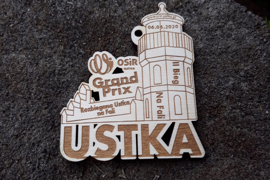 Drewniany medal na Grand Prix Ustki z precyzyjnym grawerem zabytkowej latarni morskiej, logotypem wydarzenia oraz ozdobną falą, wykonany z jasnej brzozy bez osmaleń.