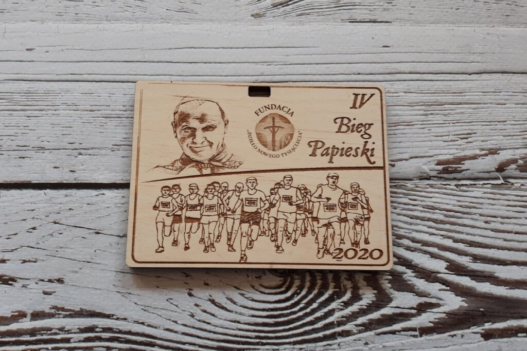 Prostokątny drewniany medal na „Bieg Papieski”, z precyzyjnym grawerem wizerunku św. Jana Pawła II, sylwetkami biegaczy oraz nazwą wydarzenia, wykonany z czystej brzozy bez osmaleń, grubość medalu 6 mm.