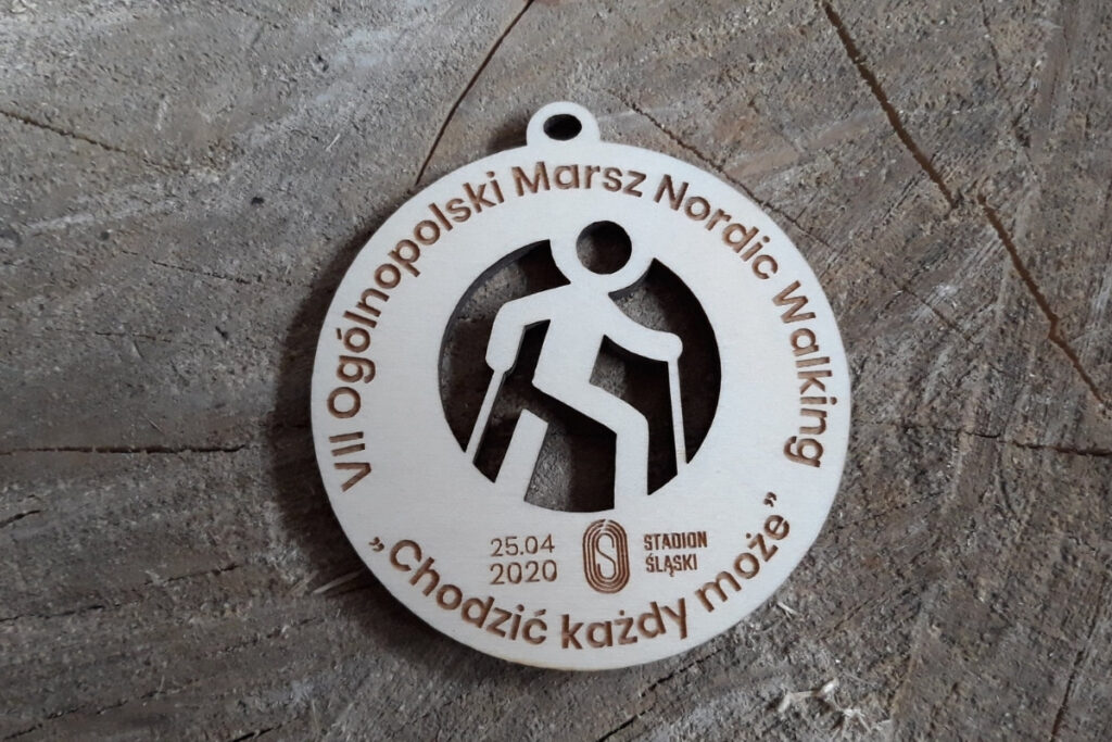 Okrągły drewniany medal na Ogólnopolski Rajd Nordic Walking „Chodzić każdy może”, z ażurowo wyciętą sylwetką osoby maszerującej z kijkami oraz grawerem logotypu wydarzenia, wykonany z czystej brzozy bez śladów dymu.