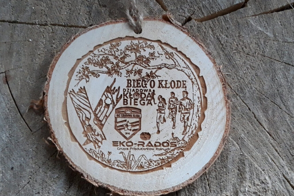 Naturalny drewniany medal na „V Bieg o Kłodę”, wykonany na autentycznym plastrze brzozy z korą, z grawerem drzewa, biegaczy oraz logotypem, zachowujący jasny odcień drewna bez osmaleń.