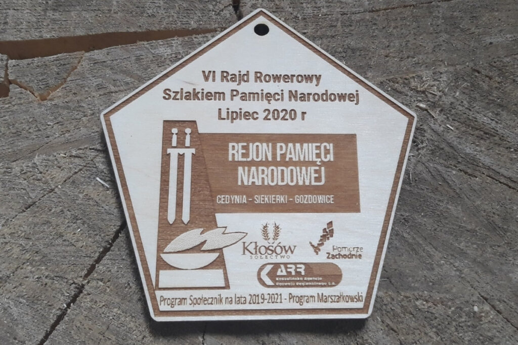 5 - kątny drewniany medal upamiętniający „Rajd Rowerowy Szlakiem Pamięci Narodowej”, z grawerem historycznych symboli,  wykonany z jasnej brzozy o nieskazitelnie czystej powierzchni bez osmaleń. Na medalu logotypy.
